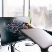 A person points at architectural blueprints displayed on a computer monitor while holding a tablet, working in a bright office with large windows in the background.