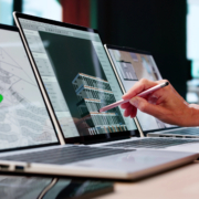 A person points at a 3D building model on a laptop screen with a pen. Two laptops beside it display architectural plans and diagrams. The workspace is modern and professional.