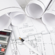 Rolled and flat architectural blueprints with technical drawings, a calculator displaying “96600,” and a black-and-white pen, all arranged on a clean white desk.
