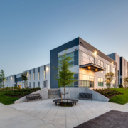 A modern industrial or office building with large windows and gray-and-white exterior, surrounded by pathways, green lawns, young trees, and benches, photographed at sunset.