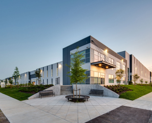 A modern industrial or office building with large windows and gray-and-white exterior, surrounded by pathways, green lawns, young trees, and benches, photographed at sunset.