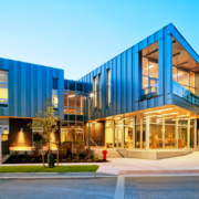 A modern building with large glass windows, metal paneling, and angular architecture is illuminated at dusk. The entrance is well-lit, and there are trees and landscaping around the exterior.