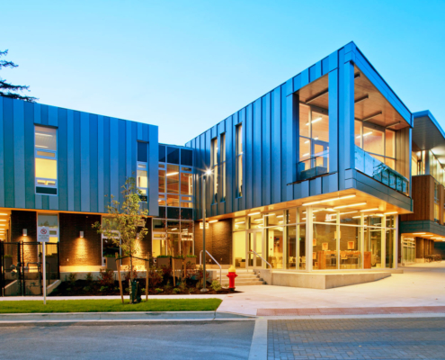 A modern building with large glass windows, metal paneling, and angular architecture is illuminated at dusk. The entrance is well-lit, and there are trees and landscaping around the exterior.