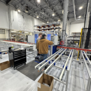 Two people operate a machine in an industrial facility with metal conveyor rails, a black tool cart, storage shelves, and high ceilings. Boxes and equipment are visible throughout the well-lit workspace.