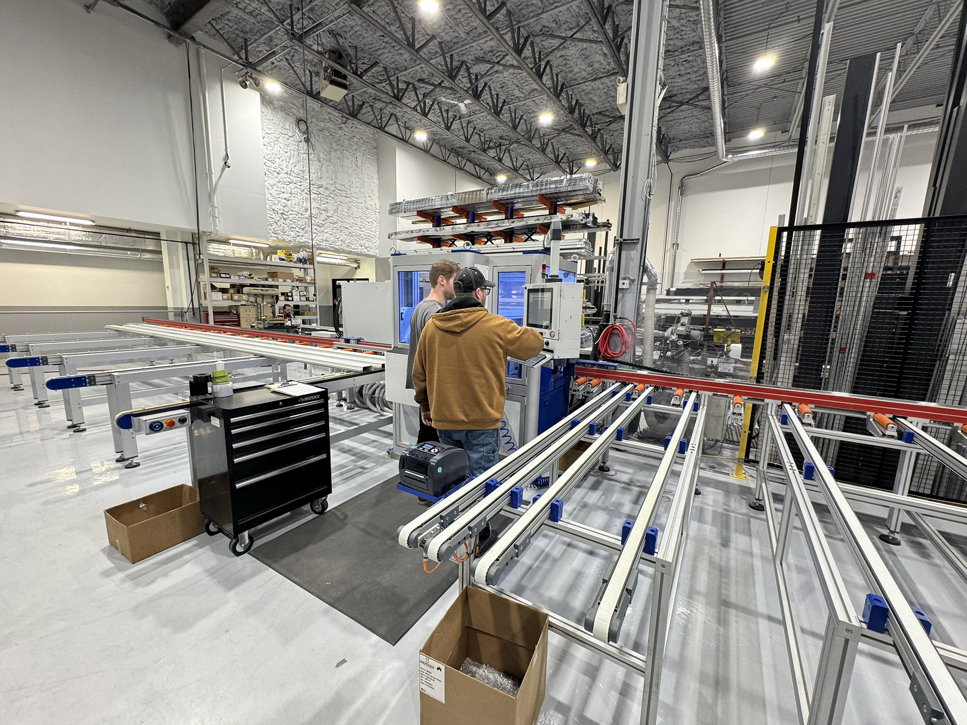 Two people operate a machine in an industrial facility with metal conveyor rails, a black tool cart, storage shelves, and high ceilings. Boxes and equipment are visible throughout the well-lit workspace.