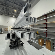 Long aluminum profiles are stacked and stored on metal racks inside a spacious warehouse with high ceilings, exposed beams, and organized shelving along the walls.