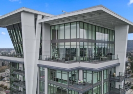 A modern high-rise apartment building with large floor-to-ceiling windows and spacious balconies, set against a clear blue sky and a cityscape in the background.