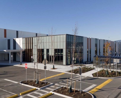 A modern industrial or warehouse building with tall windows, striped facade, and an adjacent covered walkway. The parking lot has clear road markings, stop signs, and young trees planted along the sidewalks.