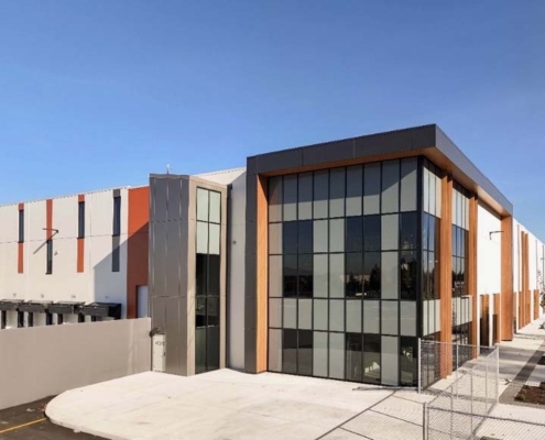 A modern industrial building with large glass windows and orange accents, featuring a loading dock area and a paved walkway with newly planted trees under a clear blue sky.