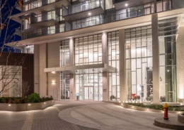 The image shows the well-lit entrance of a modern high-rise building at night, featuring large glass windows, tall white columns, and a circular driveway with landscaped areas and a yellow fire hydrant.