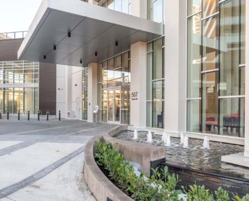 Modern building entrance with large glass windows, tall columns, and a covered walkway. In front, there is a curved planter with green plants and a small fountain beside the sidewalk.