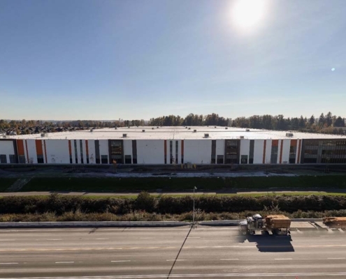 A large, modern warehouse-style building with white and orange accents stands behind a road with a truck and trailers. The sun is shining brightly in the sky, and there are trees and houses in the background.