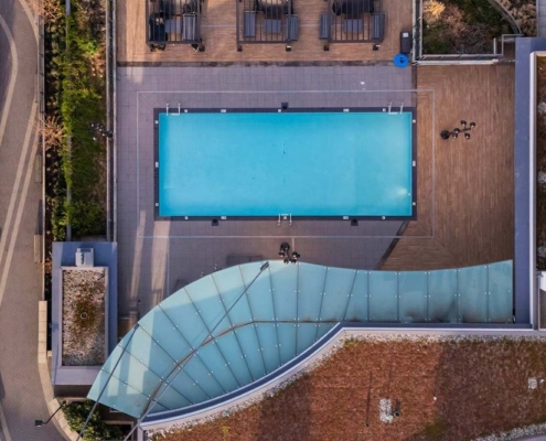 Aerial view of a rectangular swimming pool on a rooftop surrounded by lounge chairs and decking, with adjacent glass canopy and modern buildings, and landscaped areas nearby.