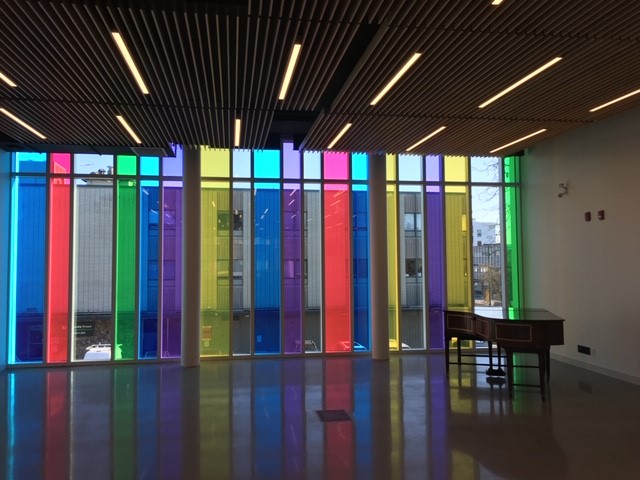 A modern room with a wooden slatted ceiling, polished floor, and tall windows covered in colorful vertical panels in red, yellow, green, blue, and purple, casting vibrant light inside. A desk sits near the windows.