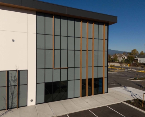 Modern building facade with large grid windows and vertical wooden beams. The structure is adjacent to an empty parking lot under clear blue sky, with trees and distant mountains in the background.