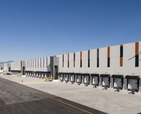 A large, modern warehouse with multiple loading docks and bay doors, set against a clear blue sky. The building has vertical orange and gray accents, and the spacious concrete area is empty except for a few vehicles in the distance.