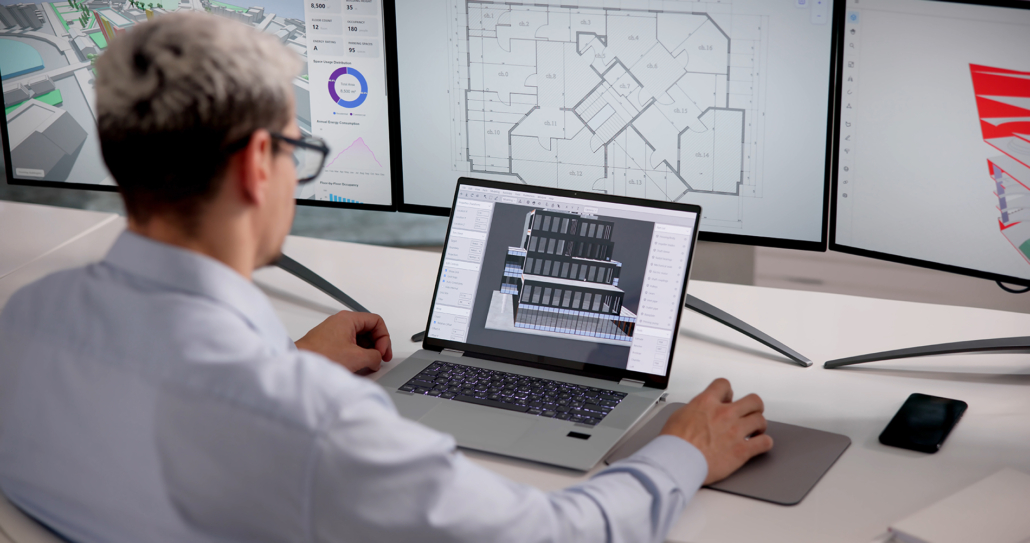 A person with short gray hair sits at a desk using a laptop with architectural software. Two large monitors display building floor plans and diagrams in the background.