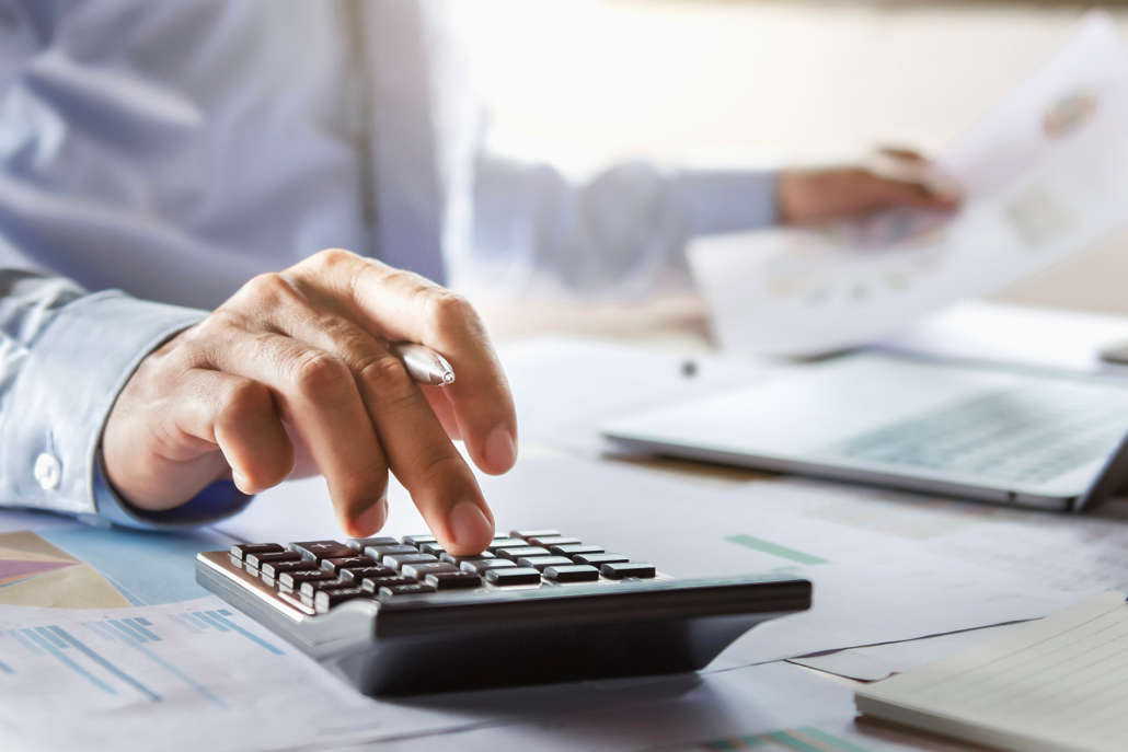 A person in business attire uses a calculator on a desk covered with documents, charts, and a laptop, suggesting financial calculations or accounting work.