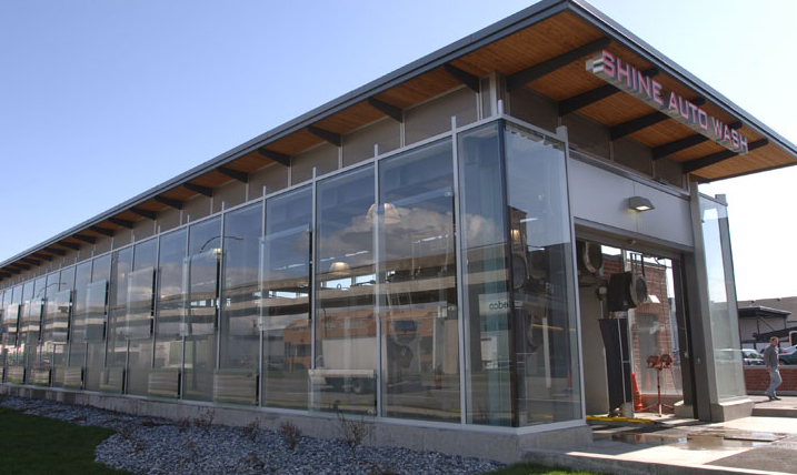 A modern glass-walled car wash building with the sign SHINE AUTOWASH on the roof, featuring a clean exterior, gravel landscaping, and a grassy area in the foreground.