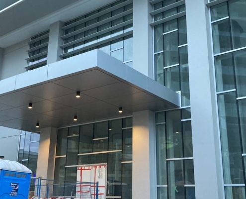 A modern building under construction with tall glass windows, a metal entrance canopy, construction materials, a blue portable toilet, and scaffolding visible in the foreground.
