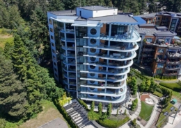 A modern, multi-story residential building with curved balconies and large windows, surrounded by trees and landscaped gardens, viewed from above on a sunny day.