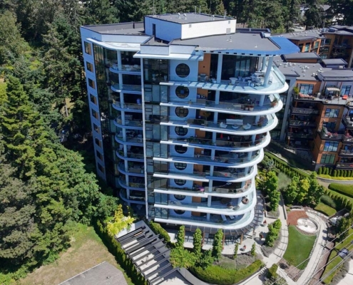 A modern, multi-story residential building with curved balconies and large windows, surrounded by trees and landscaped gardens, viewed from above on a sunny day.