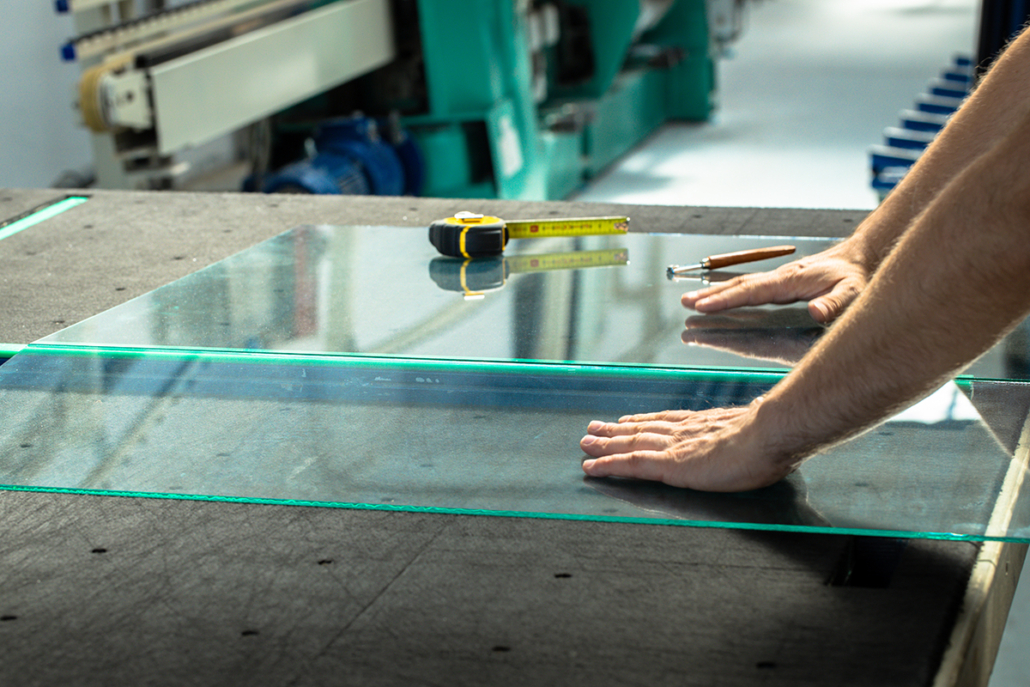 A person’s hands position a large glass sheet on a worktable, alongside a tape measure and a glass cutting tool in a factory or workshop setting.