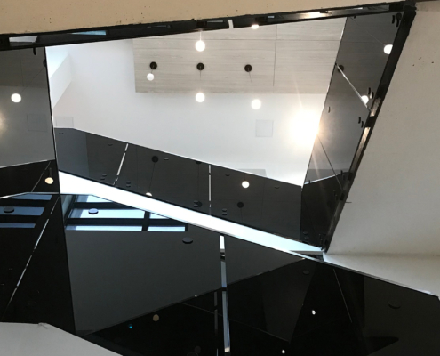 View looking up at a modern interior staircase with black glass railings, angular lines, and hanging ceiling lights. Bright natural and artificial light illuminate the minimalist architecture.