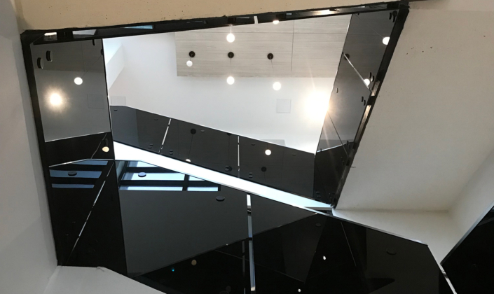 View looking up at a modern interior staircase with black glass railings, angular lines, and hanging ceiling lights. Bright natural and artificial light illuminate the minimalist architecture.