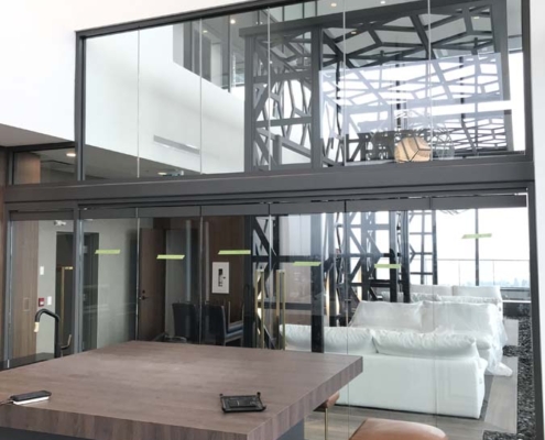 Modern, open-concept living space featuring a kitchen island with brown stools, a glass wall, white sectional sofa, and large windows letting in natural light. Contemporary decor and architectural details are visible.