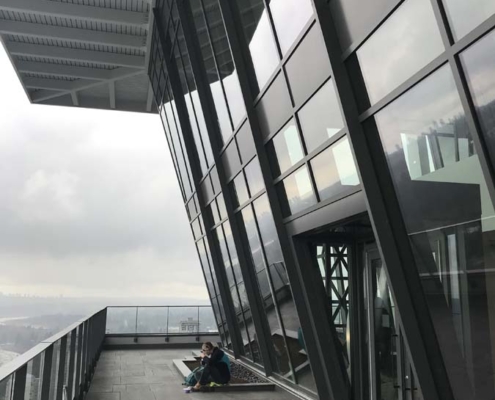A person sits on the floor near large slanted glass windows on a high-rise balcony, with outdoor furniture and equipment nearby, overlooking a cloudy cityscape.