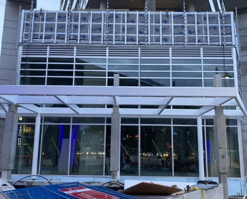 A construction site in front of a modern glass building with metal framework, unfinished pillars, blue panels, and construction materials scattered on the ground. The buildings lower levels have large glass windows and metal railings.