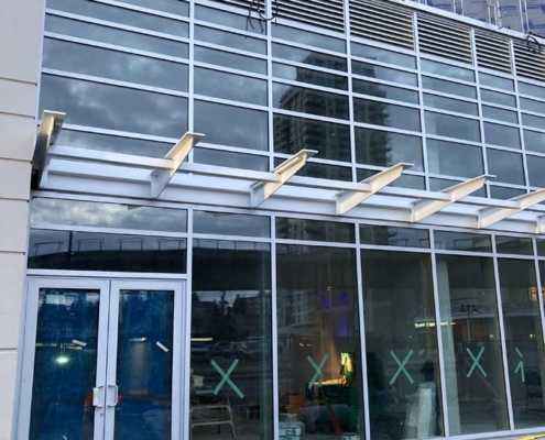 A modern glass building under construction with blue doors, tape Xs on the windows, orange traffic cones, and caution tape blocking the entrance. Building materials and a tall reflection are visible in the glass.