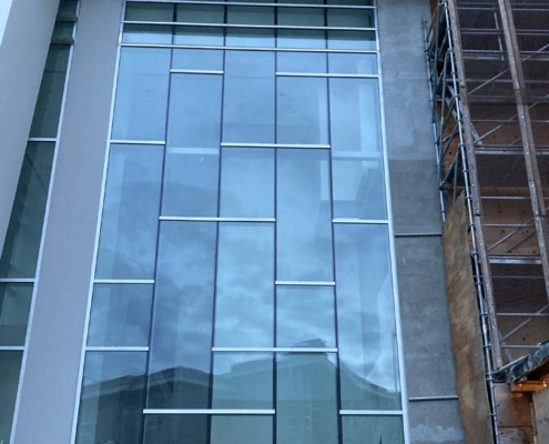 Tall modern glass building facade with large reflective windows, surrounded by unfinished concrete and scaffolding, showing signs of ongoing construction work.
