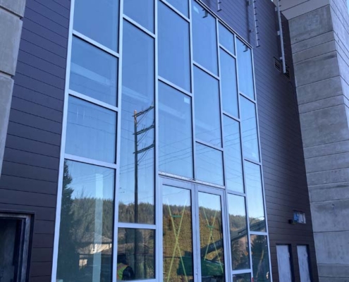 A modern building facade with tall, rectangular glass windows and white frames. Construction materials are stacked at the base, and some window panels reflect nearby trees and power lines.