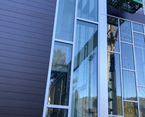 Tall, narrow glass windows set in a modern building facade with dark horizontal panels, reflecting trees, sky, and construction equipment. A green lift is visible on the right side near the upper windows.