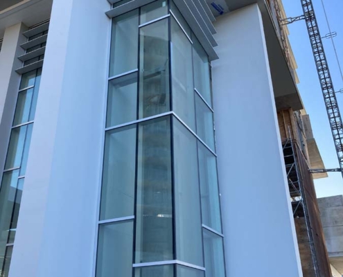 A modern building facade with tall, floor-to-ceiling glass windows framed by white walls and horizontal metal louvers above, with a construction crane visible in the background.