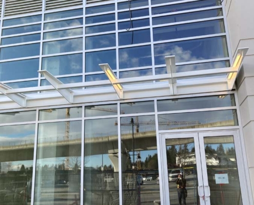 A modern building with large glass windows and double glass doors. A small overhang with metal beams extends above the entrance. The reflection shows an overpass, trees, and a blue sky with clouds.