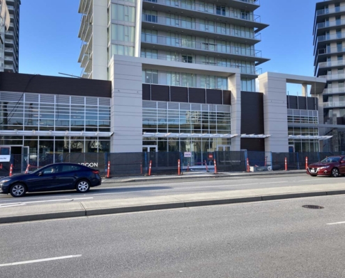 A modern, multi-story building under construction with glass windows and a fenced-off area in front. Two cars are parked on the street, and the sky is clear and blue.