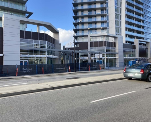 A modern high-rise building under construction with blue fencing around the site, orange traffic cones, and a green car parked on the street in front. The sign on the fence reads Building for life.