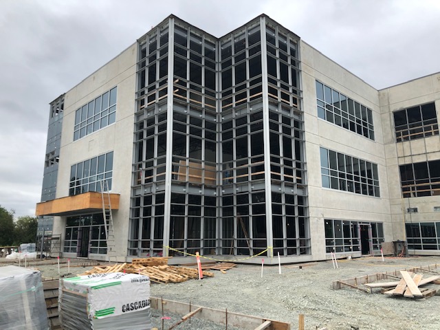 A three-story building under construction with exposed steel framework, unfinished walls, and scattered construction materials on the ground in front. The sky is overcast.
