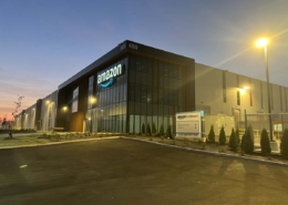 Amazon fulfillment center building at dusk, with illuminated Amazon signage on glass facade, exterior lights on, and a sign reading Amazon Fulfillment near the entrance. The sky is dimly lit, and the parking lot appears empty.