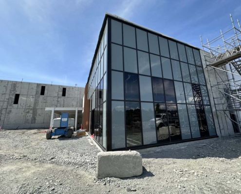 A modern building under construction with large glass windows, scaffolding on the right, a concrete wall on the left, and a blue lift on a gravel-covered ground under a clear blue sky.