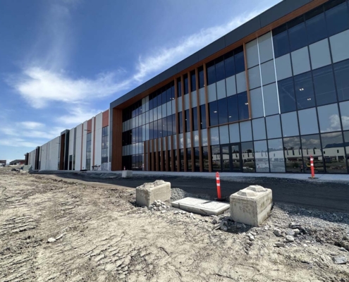 A modern, glass-fronted commercial building under construction, with exposed dirt and construction barriers in the foreground under a partly cloudy blue sky.