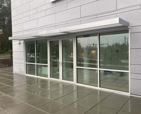 Glass double doors and large windows on a modern gray building, with a metal awning above. The wet pavement and reflections indicate recent rain. Trees and traffic lights are visible outside in the background.