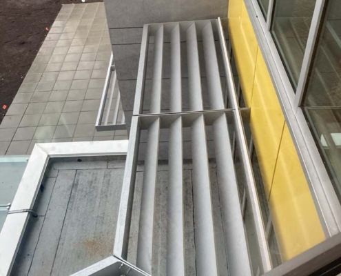 View from above of a building facade with large metal sunshades attached to yellow and gray exterior walls, above a tiled sidewalk. A person stands near the street in the background.