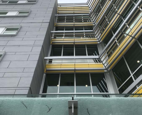 Upward view of a modern building with gray and yellow horizontal accents, large glass windows, and a green-tinted glass canopy at the entrance under a cloudy sky.