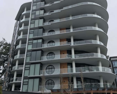 A modern multi-story apartment building with curved balconies, large windows, and a distinctive rooftop overhang, set against a cloudy sky and surrounded by greenery.