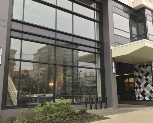 A modern building with large glass windows, a covered entrance, and a row of bike racks in front. Reflections of other buildings are visible in the windows. There are small plants and a sidewalk in the foreground.