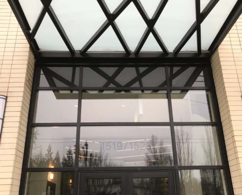 Glass building entrance with double doors and a modern black metal canopy above. Reflections of trees and cars are visible in the doors and windows. Signs and a keypad are posted near the entrance.